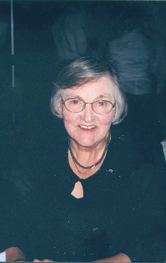 Smiling person with short gray hair and glasses, wearing a black top against a dark background