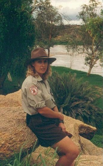 Person in a brown hat sits on a rock by a river, with greenery and trees in the background.