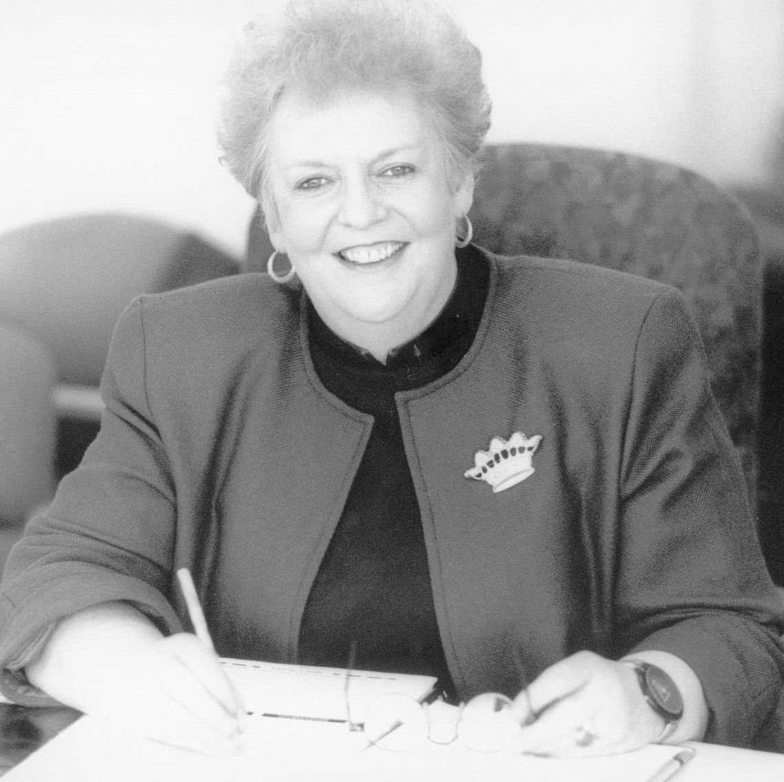Smiling person seated at a table, writing on paper in a black-and-white photo