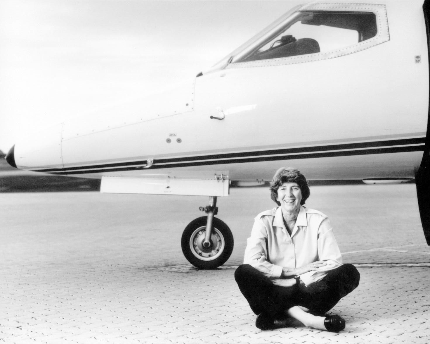Person sitting cross-legged beside a small airplane on a tarmac, smiling at the camera.