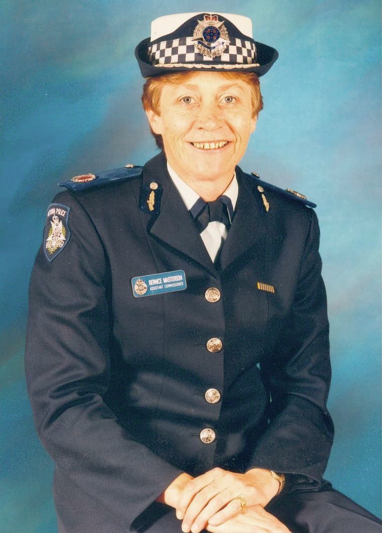 Police officer in dark uniform and checkered cap, seated and smiling against a blue studio backdrop.