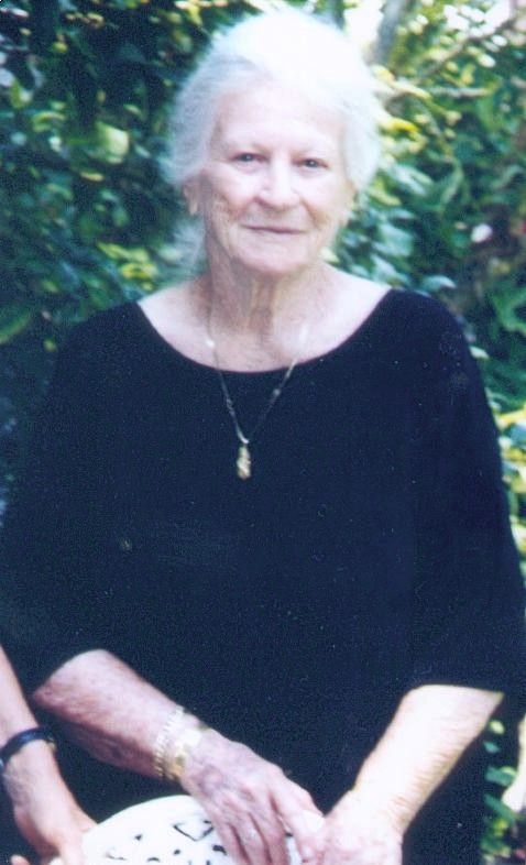 Elderly woman in a black top sitting outdoors against leafy greenery, wearing a small gold pendant.
