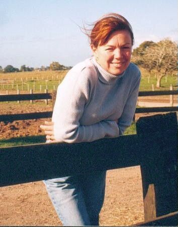 Person leaning on a wooden fence in a sunny field, wearing a light sweater and jeans.