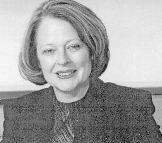 Portrait of a smiling person with short hair, wearing a dark jacket and patterned top, against a plain background