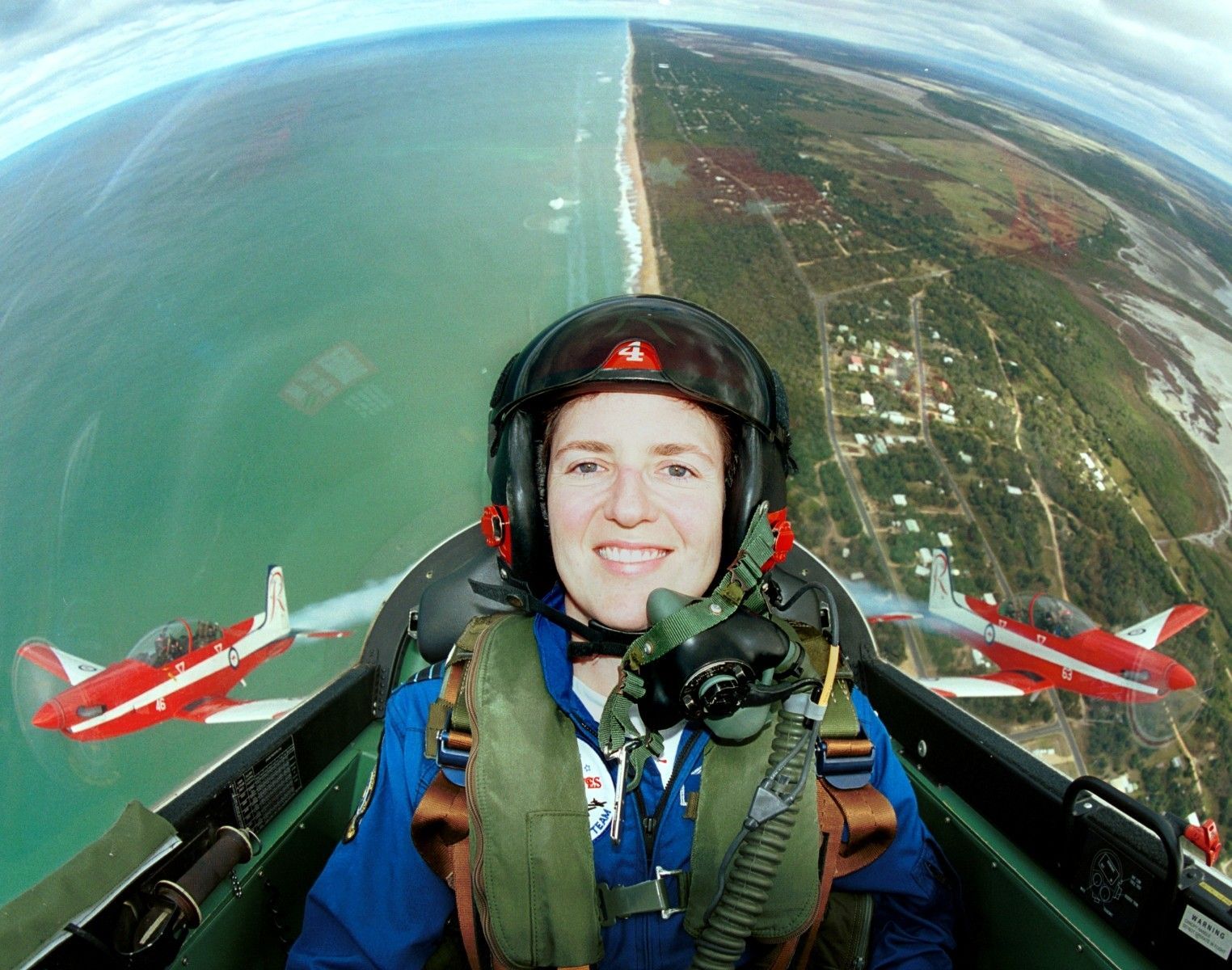 Pilot smiling in a cockpit over a coastline, with two red jets flying nearby.