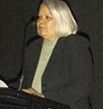 Woman with long platinum blonde hair in a dark blazer speaking at a podium against a black background