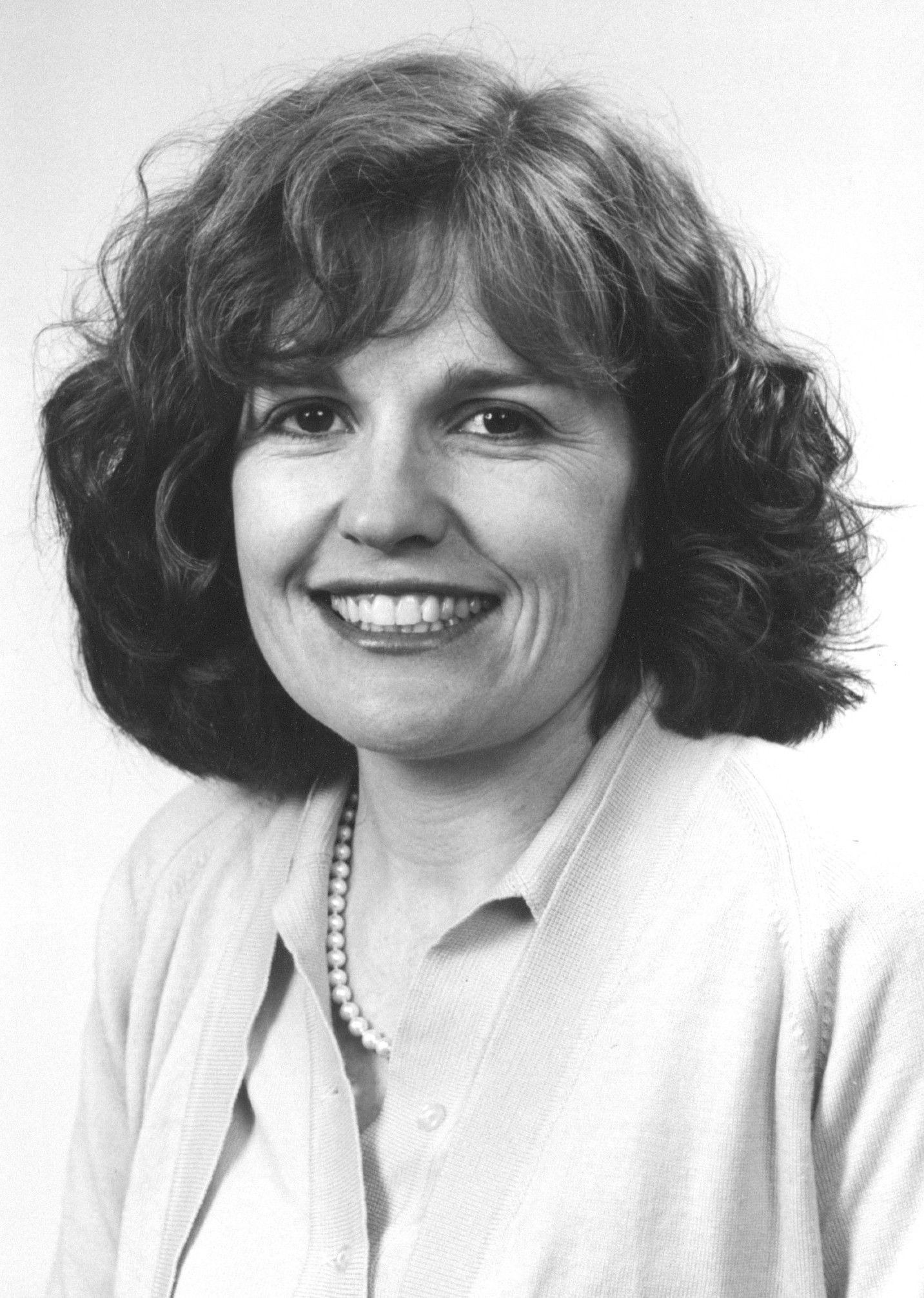 Black-and-white portrait of a smiling woman with curly hair, wearing a light blouse and necklace.