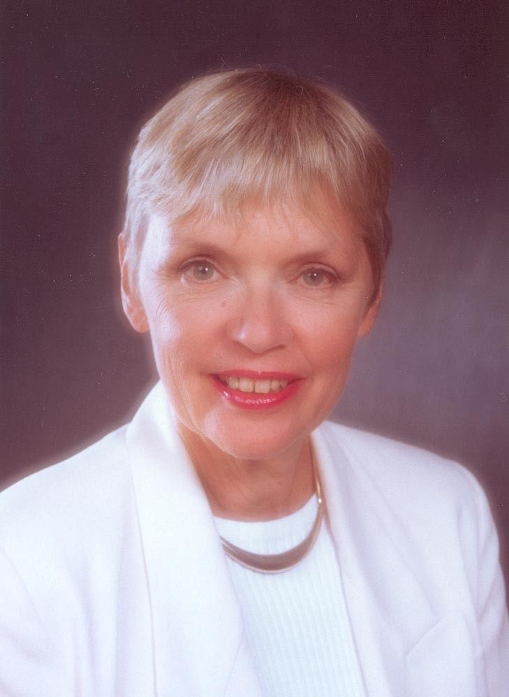 Studio headshot of a smiling woman with short blonde hair in a white blazer and necklace on a gray background