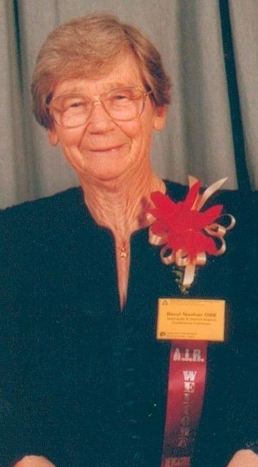 Elderly woman in glasses wearing a dark jacket and a red floral medal ribbon, posing indoors.