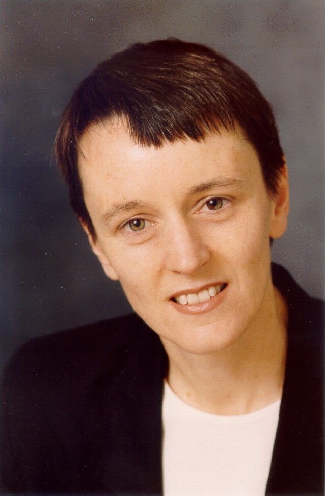Portrait of a person with short dark hair, wearing a black jacket and white shirt, against a gray background