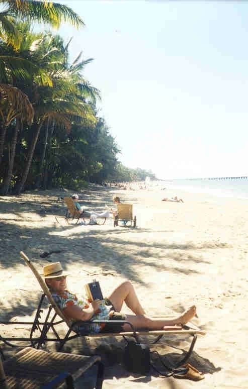 People relaxing on lounge chairs along a sunny tropical beach with palm trees and calm water.