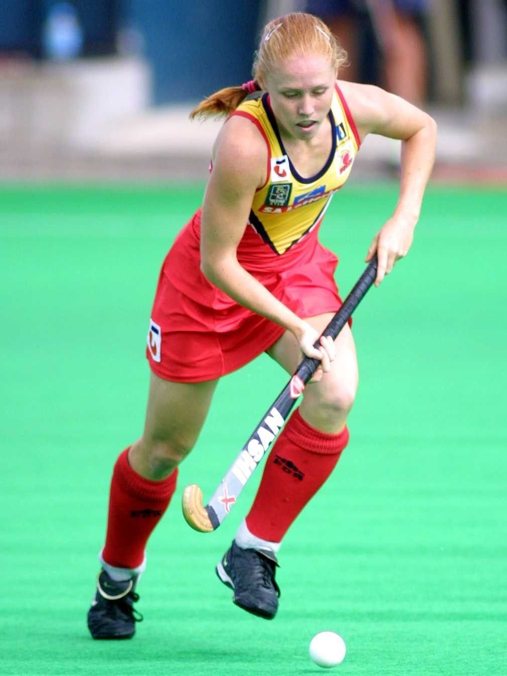 Field hockey player in red and yellow uniform dribbling a white ball on turf