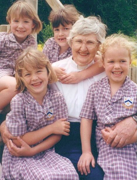 Woman seated outdoors with four children in matching plaid uniforms, smiling at the camera.