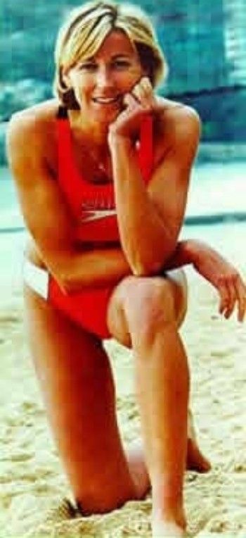 Woman in a red swimsuit kneeling on a sandy beach, smiling at the camera.