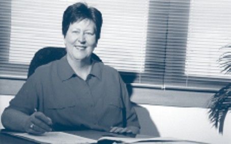 Person sitting at a desk, smiling and holding a pen in a sunlit office.