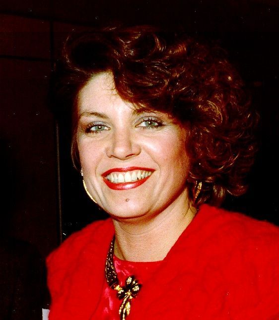 Smiling woman in a red jacket with curly dark hair and red lipstick against a dark background