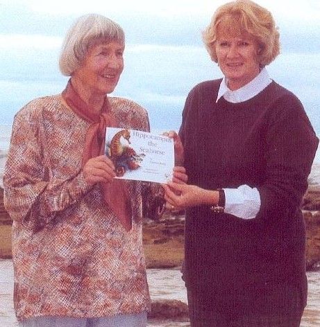 Two women standing outdoors, smiling while holding a certificate against a cloudy sky.