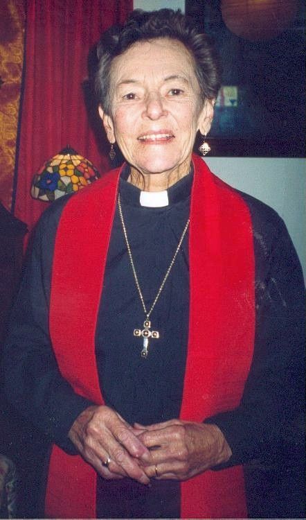 Clergy member in black robe with red stole and cross, standing indoors and smiling