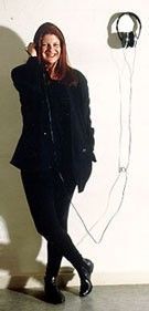 Person in a black outfit leaning against a white wall beside hanging headphones.