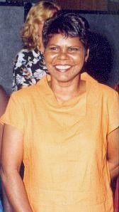 Smiling person in an orange shirt standing indoors, with another person blurred in the background