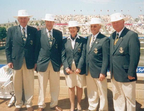 Five people in white hats and navy blazers stand together on a sunny boardwalk overlooking a waterfront.
