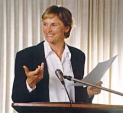 Speaker at podium holding papers and gesturing, with a microphone in front of a striped backdrop