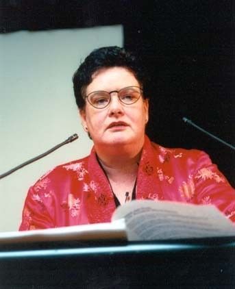 Speaker at a podium in a red jacket, speaking into microphones with papers in front of her