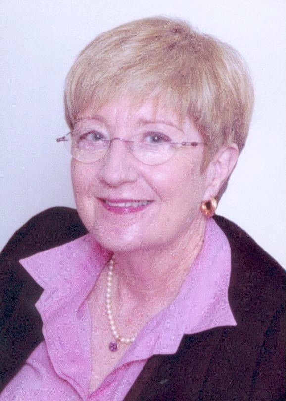 Smiling woman with short blonde hair, glasses, and a pink blouse under a dark jacket.