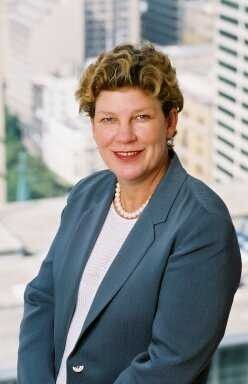 Smiling woman in a blue blazer with a pearl necklace, standing before a blurred cityscape background.