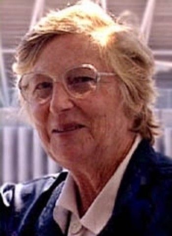 Portrait of a smiling person with glasses and short blond hair in a dark jacket and white collared shirt