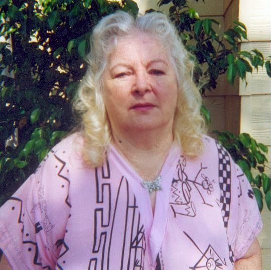 Woman with long blonde hair in a pink patterned shirt standing outdoors by leafy greenery