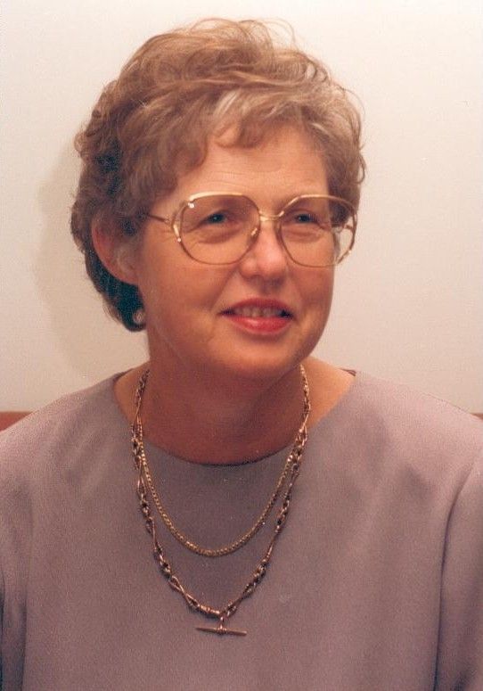 Portrait of a woman with glasses and short curly hair, wearing a lavender top and layered necklaces.