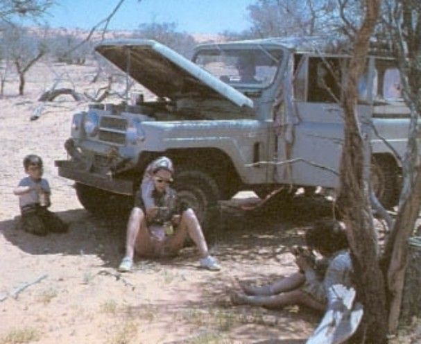 Three people sit by a white off-road vehicle with its hood open in a dry, wooded area.