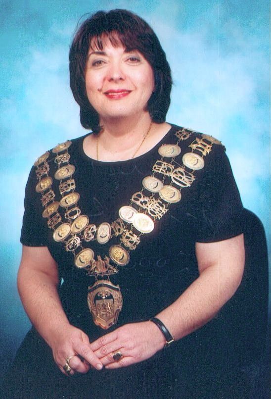 Woman in black dress wearing a large ceremonial medallion sash against a blue backdrop