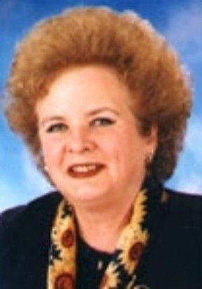 Head-and-shoulders portrait of a smiling person in a dark jacket and patterned blouse against a blue background