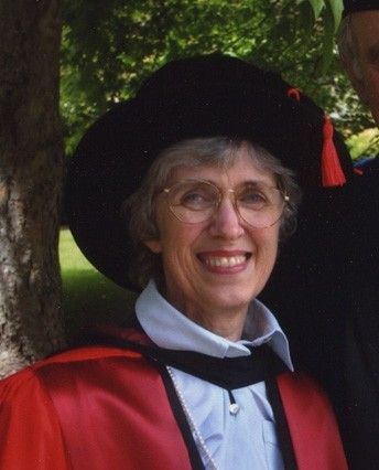 Smiling woman in graduation cap and red gown standing outdoors by a tree.