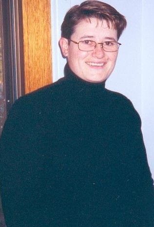 Person smiling in a dark turtleneck sweater, standing indoors by a wooden door.