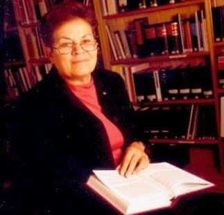 Woman seated at a library table with an open book, shelves of books behind her.