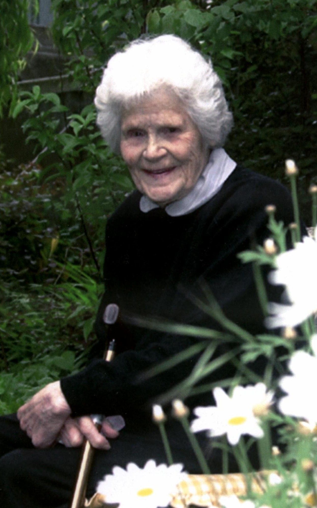 An elderly woman with white hair sits smiling in a garden among white flowers.