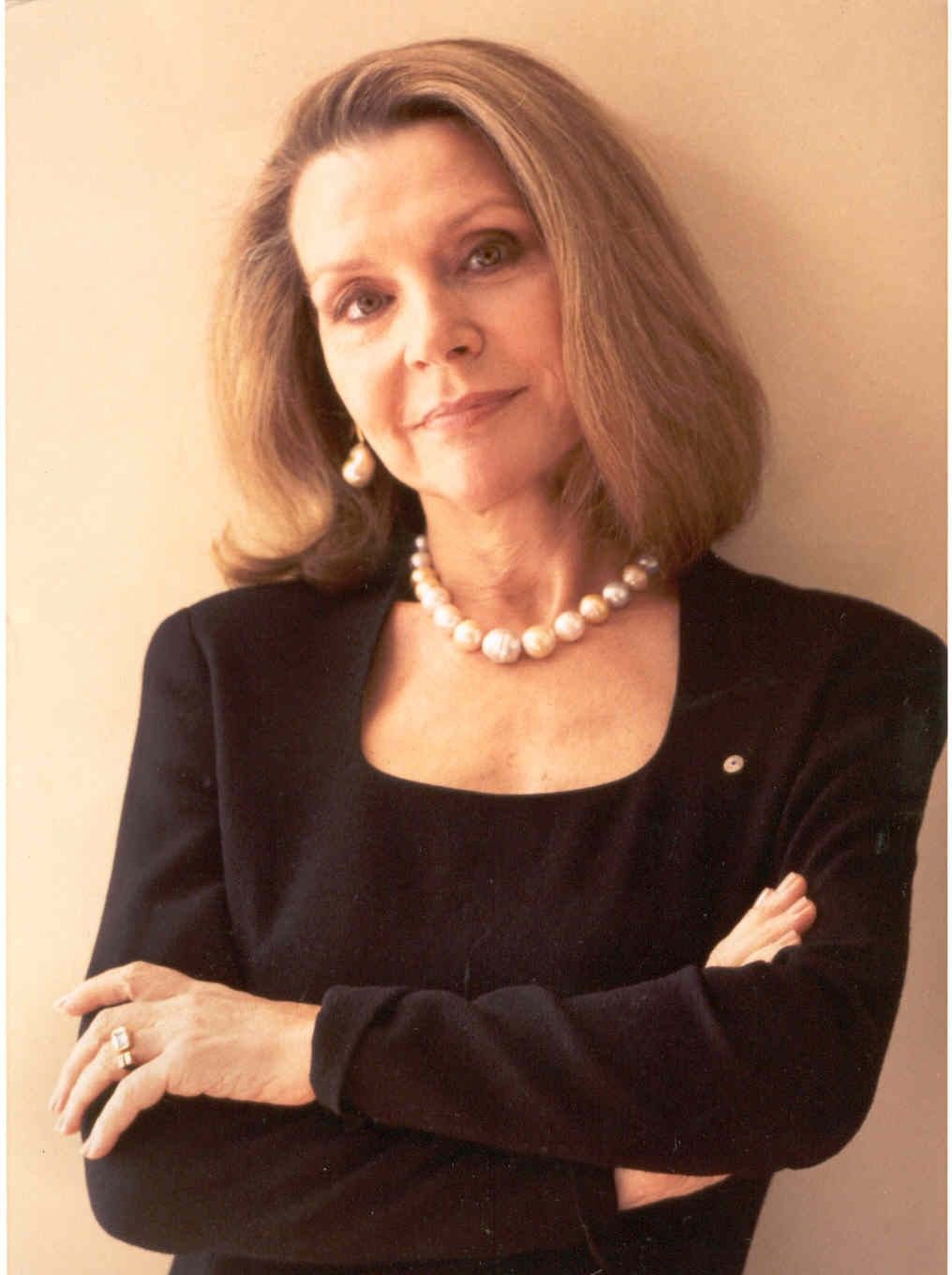 Woman in a black top with pearl necklace, standing with arms crossed against a warm beige background.