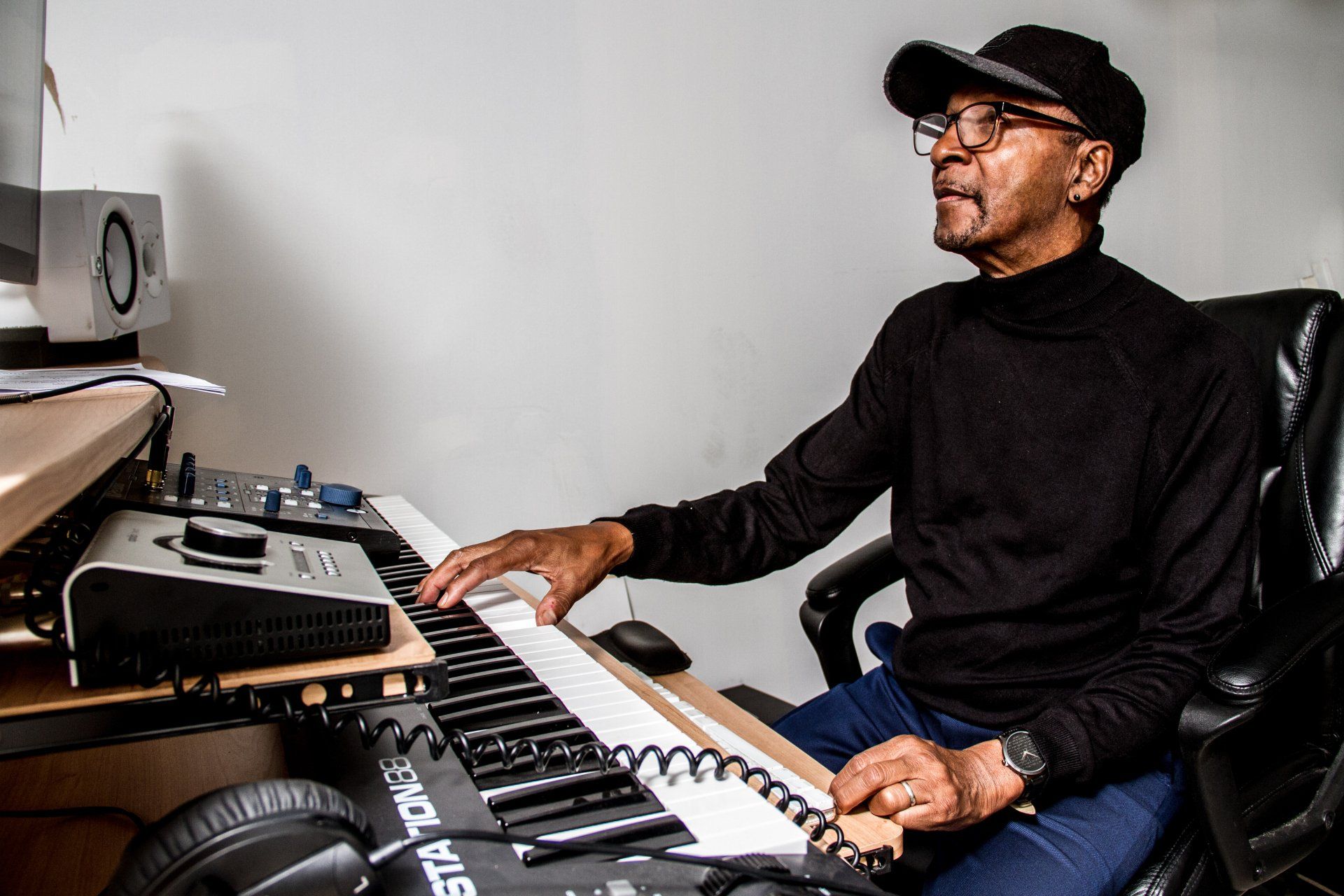 Musician in a black cap and shirt playing keyboard in a home studio, facing a computer monitor