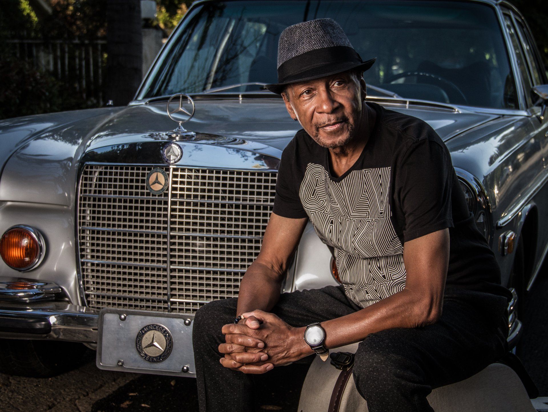 Man sitting in front of a classic black car, wearing a hat and patterned shirt.