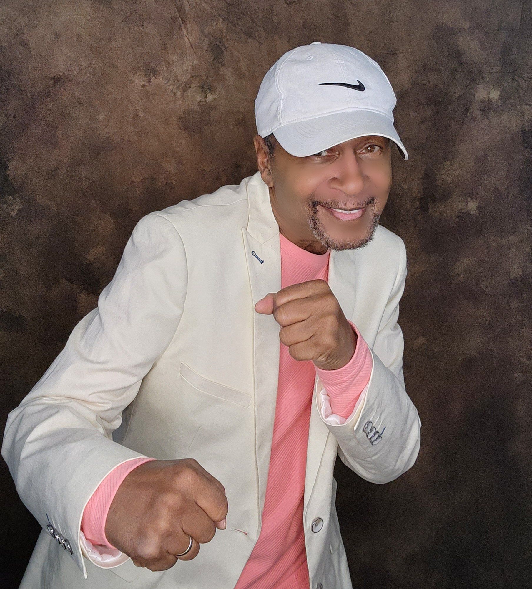 Person posing with fists up, wearing a white cap, white jacket, and pink shirt against a dark backdrop