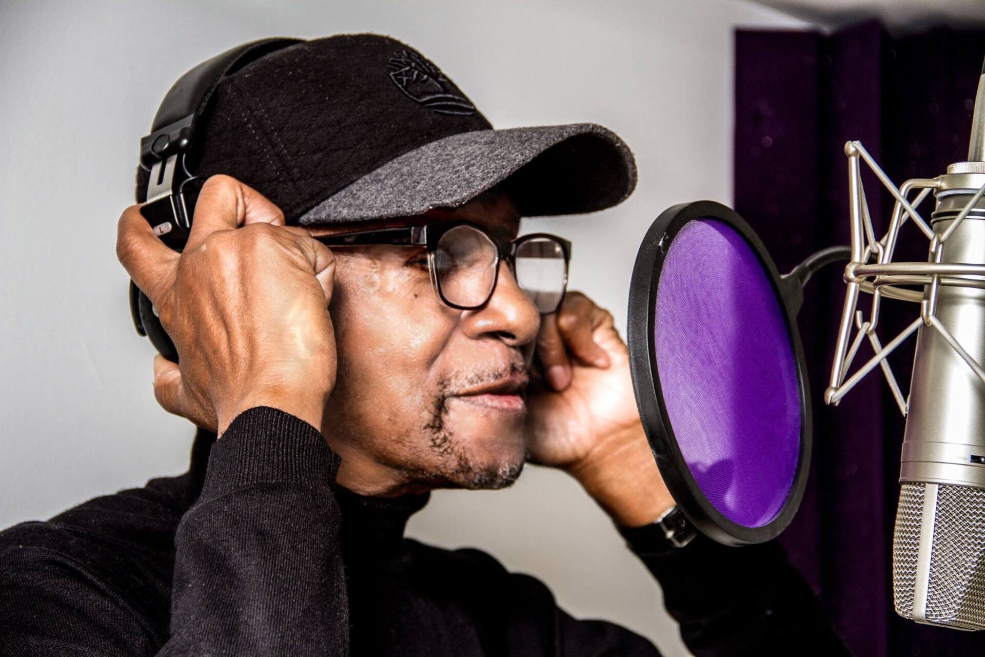 Person wearing headphones and cap singing into a studio microphone behind a purple pop filter