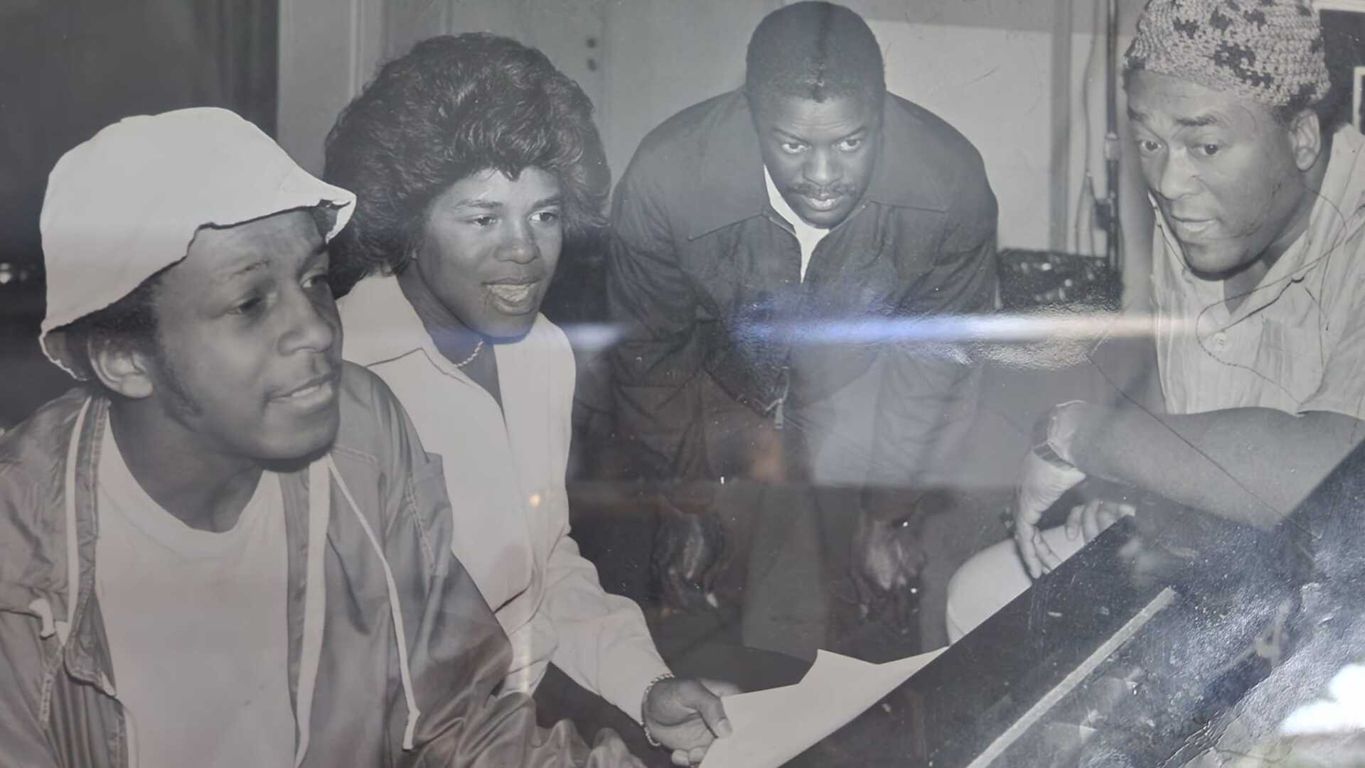 Four people gathered around an audio console, leaning in and looking down at the controls.