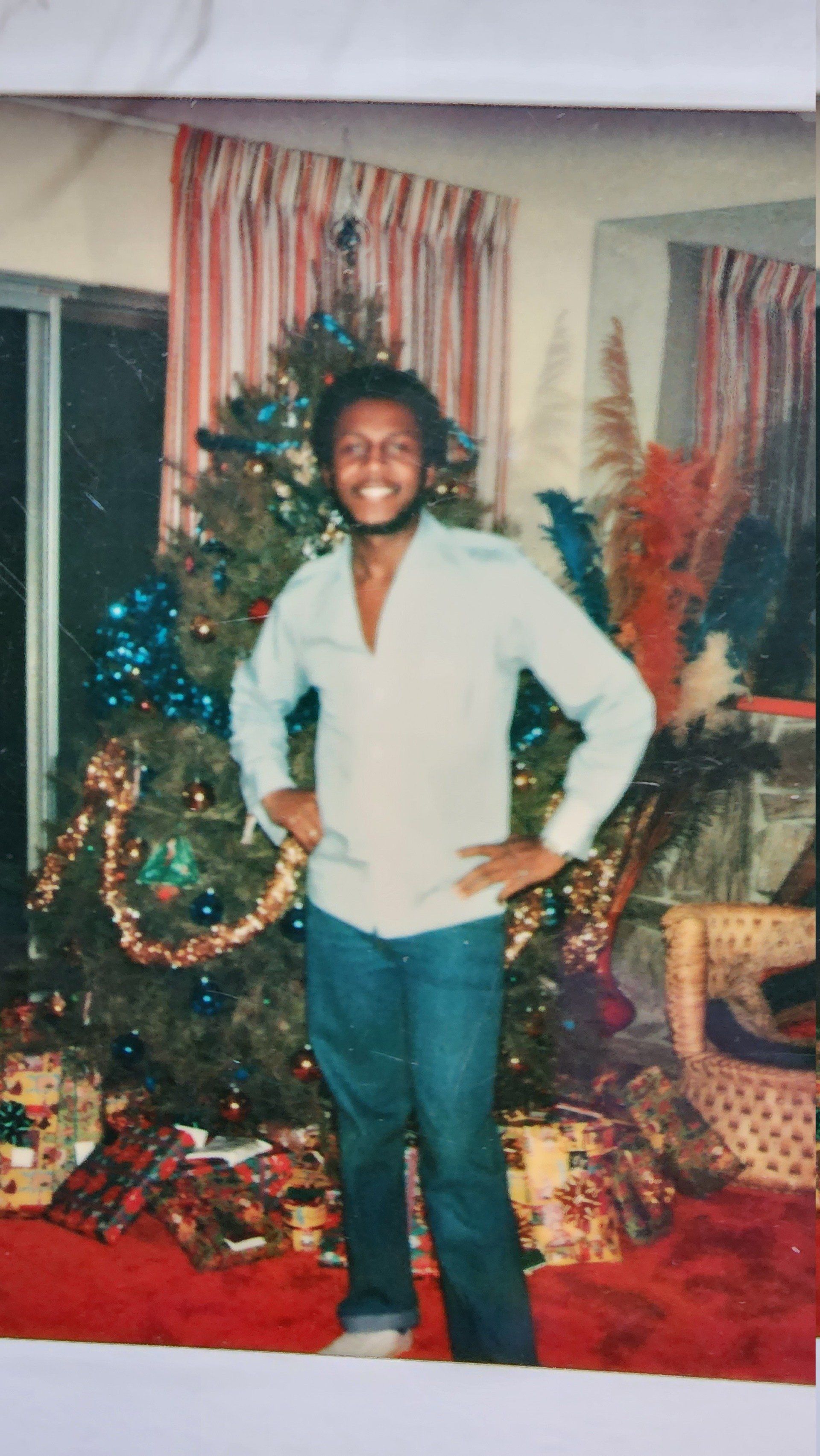 Person standing in front of a decorated Christmas tree and wrapped gifts, smiling with hands on hips