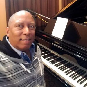 Man smiling beside a black grand piano with sheet music indoors