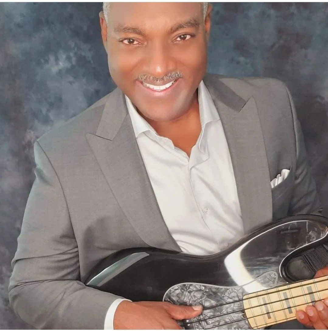 Smiling person in a gray suit holding a black electric guitar against a gray backdrop