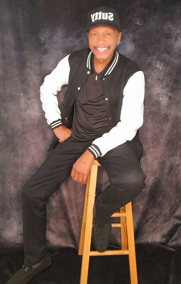 Person seated on a stool against a dark backdrop, smiling and wearing a black cap and varsity jacket.
