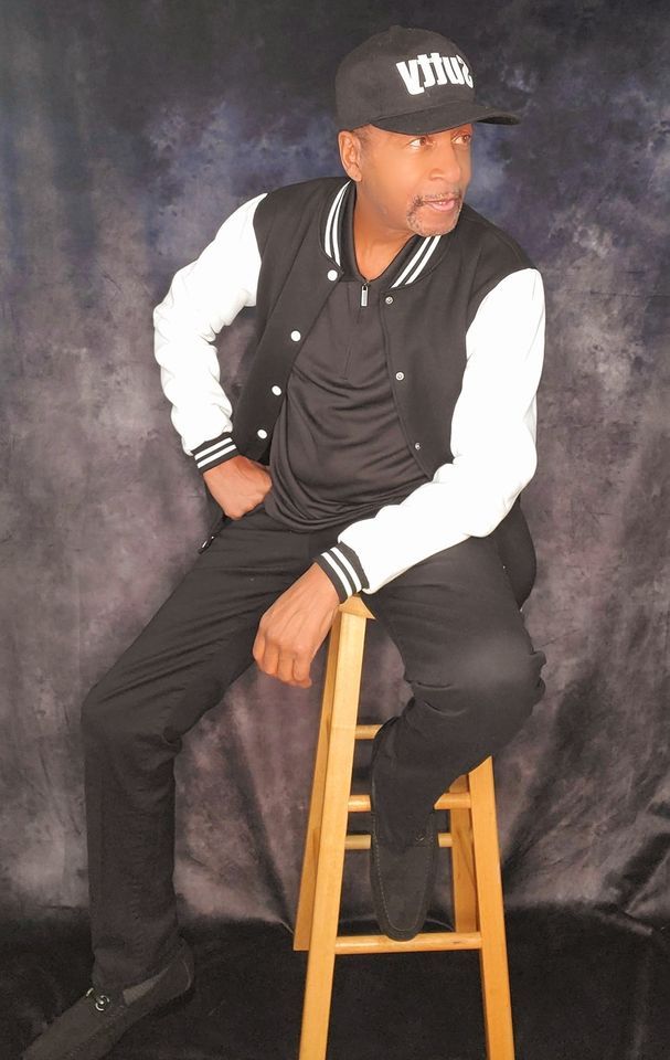 Man seated on a wooden stool in a studio, wearing a black-and-white varsity jacket and cap.